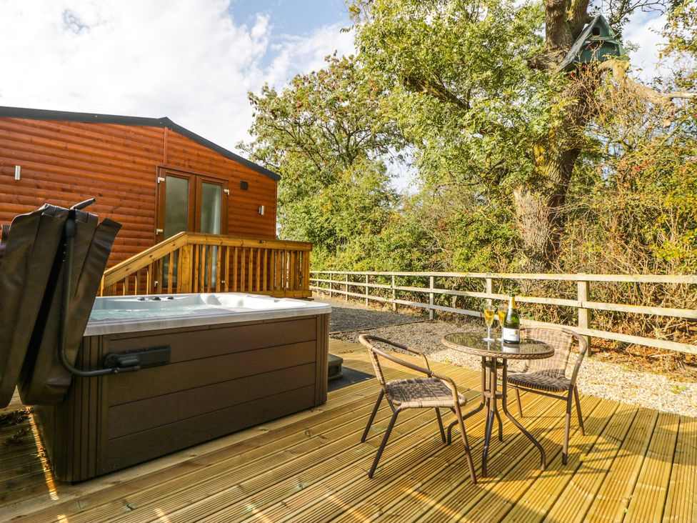 An outdoor wooden deck with a hot tub two wicker chairs and a glass table with a bottle and glasses near a wooden cabin at Beech Tree Lodge in Stamford Bridge