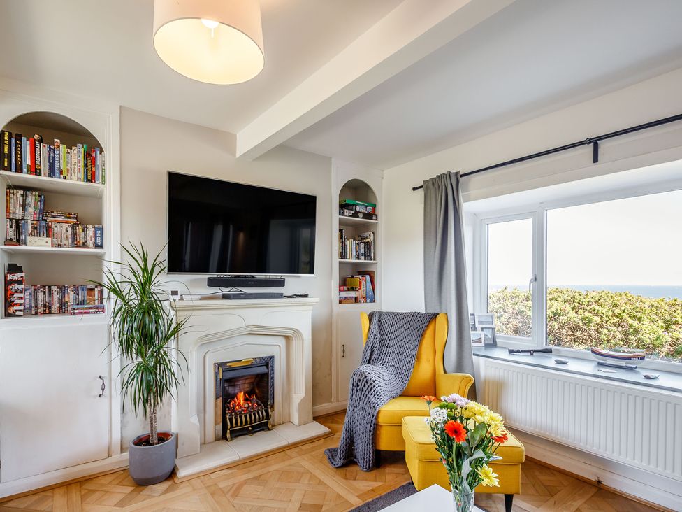A living room with a yellow armchair and ottoman next to a window with grey curtains a lit fireplace with a TV mounted above and bookshelves on either side at Tyn Y Berth in Benllech