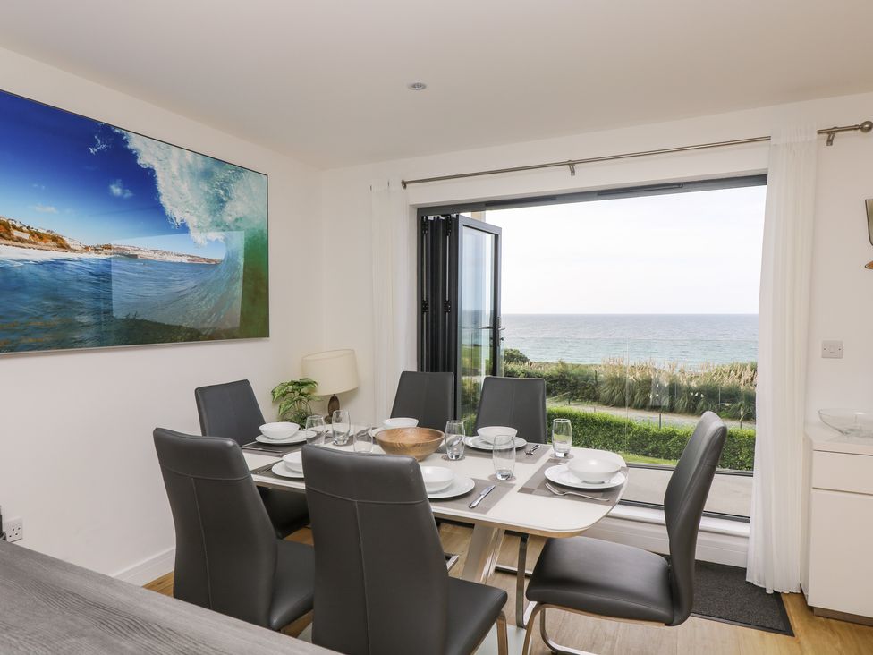 A dining area with a table set for six by a large window overlooking the sea at Praa Waves in Praa Sands